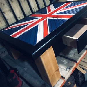 May include: A coffee table with a drawer, featuring a Union Jack design painted on the top. The table has black legs and a black frame around the top. The legs are made of natural wood.