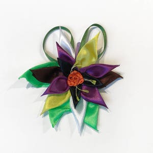 Puede incluir: Un accesorio floral para el cabello hecho a mano con cintas de satén de colores. La flor presenta tonos de morado, verde, amarillo y marrón, con un pequeño centro naranja. Las cintas están dispuestas para parecerse a pétalos y hojas.