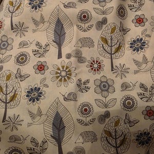 Puede incluir: Tela color crema con un patrón repetido de árboles estilizados, flores, pájaros, caracoles, mariposas y erizos. El diseño presenta una paleta de azul, rojo, marrón y gris sobre un fondo neutro. El tema general es la naturaleza y el aire libre.