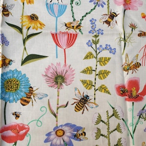 Puede incluir: Una tela con un patrón repetido de abejas y flores coloridas. Las flores incluyen girasoles amarillos, margaritas azules, cosmos rosas y amapolas rojas. Las abejas son negras y amarillas, sobre un fondo blanquecino.