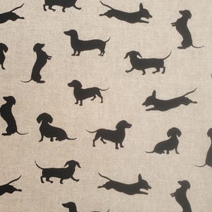 Puede incluir: Una tela beige con un patrón repetido de siluetas negras de perros salchicha en varias poses. Los perros se representan de pie, sentados y corriendo por la superficie.
