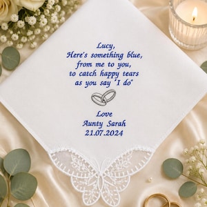 Puede incluir: Un pañuelo cuadrado blanco con texto bordado en azul que dice: Lucy, Here's something blue, from me to you, to catch happy tears as you say 'I do'. Debajo del texto hay un icono de anillos bordado en gris, seguido de Love, Aunty Sarah y 21.07.2024. El borde inferior tiene un encaje blanco con forma de mariposa.