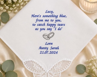May include: A white square handkerchief featuring blue embroidered text that reads: Lucy, Here's something blue, from me to you, to catch happy tears as you say 'I do'. Below the text is a grey embroidered ring icon, followed by Love, Aunty Sarah, and 21.07.2024. The bottom edge has a white lace butterfly trim.
