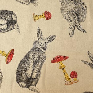 May include: A light beige fabric featuring a repeating pattern of black and white rabbits in various poses, interspersed with small yellow-stemmed mushrooms that have bright red caps with white spots, creating a detailed, nature-inspired textile design.