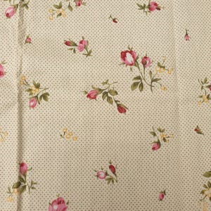 Puede incluir: Tela de color crema con un patrón repetido de pequeñas rosas rosas, hojas verdes y diminutos racimos de flores amarillas. El fondo presenta un sutil diseño de lunares en color marrón apagado. La tela tiene una estética floral vintage.