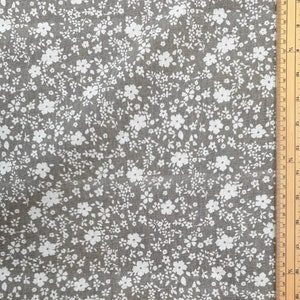 Può includere: Tessuto grigio con motivo floreale bianco. Il tessuto presenta una fitta disposizione di piccoli fiori bianchi e foglie delicate su sfondo grigio. Un righello di legno con misure in centimetri è visibile sul bordo destro.