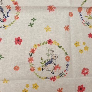 May include: Off-white fabric with a repeating pattern of bunnies inside floral wreaths. The wreaths feature red, orange, and yellow flowers with green leaves. Scattered around are small flowers in yellow, red, and orange.