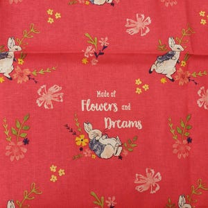 Puede incluir: Una tela roja con un patrón repetido de conejos, flores y las palabras "Dreams" y "Made of Flowers and Dreams." Los conejos son blancos con detalles azules y grises, y las flores son rosas, amarillas y blancas.
