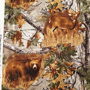 Puede incluir: Tela de camuflaje con un diseño de ciervo y oso en un entorno forestal. La tela presenta tonos marrones, verdes y blancos y el logotipo de Realtree. Medidas en centímetros.