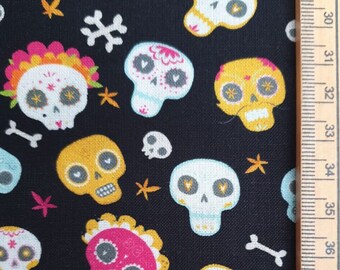 May include: Black fabric with a repeating pattern of colorful sugar skulls, bones, and star shapes. The skulls are in shades of pink, yellow, and white, with floral and decorative details. A ruler is on the right side.