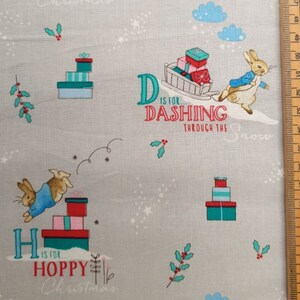 May include: A light gray fabric featuring a Christmas-themed design. The pattern includes illustrations of a rabbit, gift boxes, holly sprigs, and text that reads "H is for Hoppy Christmas" and "D is for Dashing Through the Snow".
