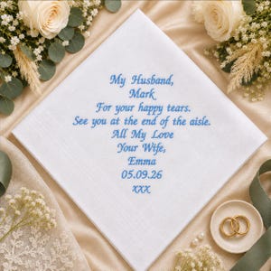 May include: A white square handkerchief featuring blue embroidered text that reads: My Husband, Mark, For your happy tears. See you at the end of the aisle. All My Love, Your Wife, Emma, 05.09.26, XXX. The fabric has a subtle border detail.