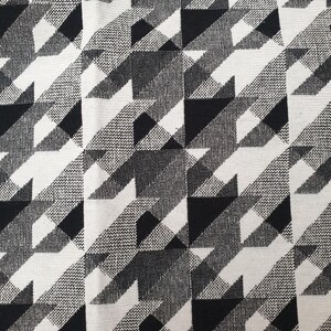 Puede incluir: Una tela tejida con un patrón repetitivo de pata de gallo en blanco y negro de alto contraste. El textil muestra un tejido denso y texturizado con formas geométricas nítidas que crean una cuadrícula entrelazada consistente en toda la superficie.