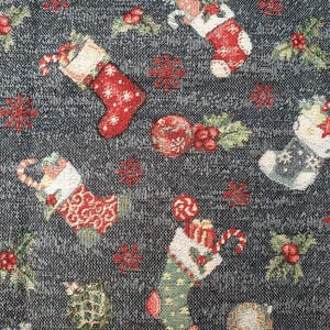 Op de afbeelding: Een donkergrijze geweven stof met een herhalend kerstpatroon van rode en groene kousen gevuld met zuurstokken en cadeautjes, afgewisseld met rode ornamenten, hulstbessen, groene bladeren en kleine rode sneeuwvlokken in het ontwerp.