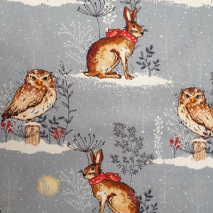 May include: A pale blue fabric with a winter design. It features brown hares with red scarves and owls on wooden posts. The pattern includes white snow, red berries, and stylized plants.