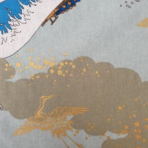 May include: A fabric with a Japanese-inspired design featuring a crane in flight, gold accents, and stylized waves in blue and white. The background is a light blue-gray color, with brown clouds.