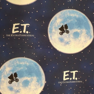 May include: Blue fabric with a repeating pattern of a white moon with a silhouette of a bicycle and a figure on it. The text "E.T. THE EXTRATERRESTRIAL" is printed in white on the fabric.