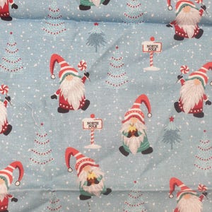 May include: Light blue fabric with a repeating pattern of Christmas gnomes, Christmas trees, and North Pole signs. The gnomes wear red hats and coats, and hold candy canes. The trees are light blue with red dot ornaments.