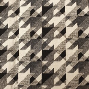 Puede incluir: Una tela tejida con un patrón repetitivo de pata de gallo en negro, blanco y tonos de gris de alto contraste. El textil muestra un tejido denso y texturizado con formas geométricas nítidas dispuestas en una cuadrícula sobre la superficie.