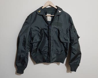 Könnte beinhalten: Eine dunkelgrüne Bomberjacke mit Reißverschluss und zwei Taschen. Die Jacke hat einen Aufnäher auf dem linken Ärmel und zwei goldene Pins am Kragen.