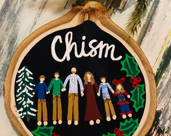 May include: A round wooden ornament with a black background. The ornament is painted with a family of six people, a Christmas tree, and holly. The text "Chism" and "2018" are painted on the ornament.