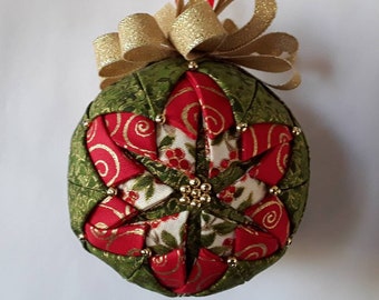 May include: A handcrafted Christmas ornament with a green and red quilted design. The ornament features a gold bow and small gold beads. The red sections have a white floral pattern. A festive holiday decoration.
