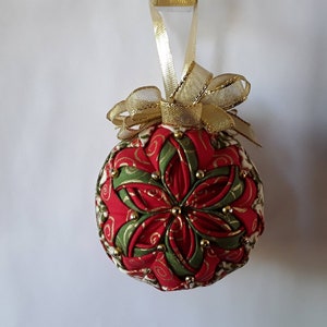 May include: A handcrafted Christmas ornament with a red, green, and gold floral design. The ornament is embellished with small gold beads and a gold ribbon for hanging. The ornament is approximately 3 inches in diameter.