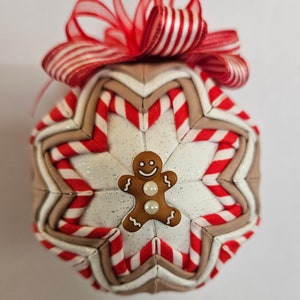 May include: A festive Christmas ornament with a starburst design. The ornament features red and white striped fabric, tan fabric, and a gingerbread man embellishment. A red and white striped bow tops the ornament.