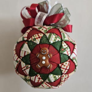 May include: A handmade Christmas ornament with a gingerbread man design. The ornament features a quilted pattern in red, green, and white fabrics. A red and white ribbon bow tops the ornament, and a small gingerbread man is centered on the front.