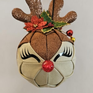 May include: A Christmas ornament shaped like a reindeer head. The ornament has brown glitter antlers, a red nose, and black eyelashes. It is decorated with a red flower and green leaves.