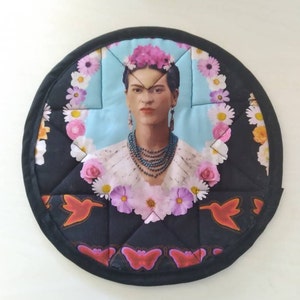 Puede incluir: Salvamanteles redondo y acolchado con un retrato de Frida Kahlo. El retrato está rodeado de un diseño floral con flores rosas, blancas y amarillas. El borde es negro con un patrón decorativo.