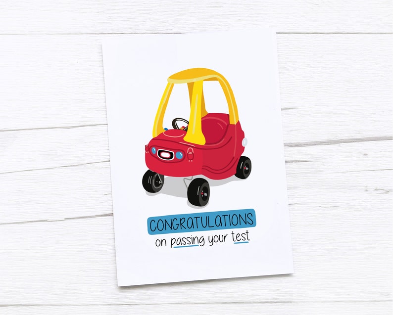 May include: A greeting card featuring a red and yellow toy car illustration. The card has the text "CONGRATULATIONS on passing your test" in a blue rectangle. The card is on a white wooden surface.