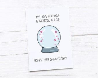 May include: A white card with a cartoon snow globe with pink hearts inside. The text on the card reads "My love for you is crystal clear" and "Happy 15th Anniversary".