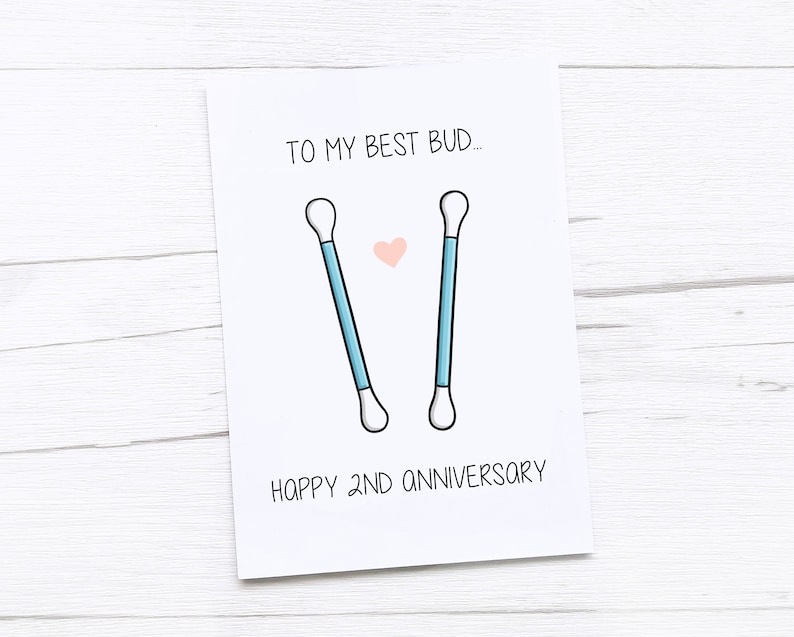 Op de afbeelding: Een witte kaart met de tekst "To my best bud..." en een tekening van twee wattenstaafjes met een roze hart ertussen. De tekst "Happy 2nd Anniversary" staat onderaan de kaart.