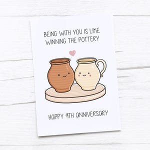 Puede incluir: Una tarjeta blanca con el texto "Estar contigo es como ganar la alfarería" y "Feliz 9º aniversario". La tarjeta presenta ilustraciones de dos vasijas de cerámica sonrientes y un corazón rosa.