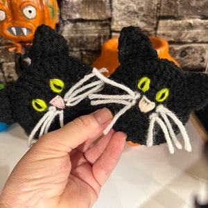 May include: Two black crocheted cat faces with white yarn whiskers and green yarn eyes. The cat faces are held in a hand.