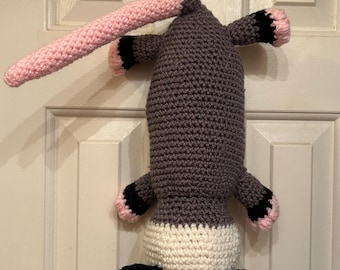 May include: A handmade crochet opossum in shades of gray, white, black, and pink. The opossum has a long pink nose, small black ears, and black feet with pink toes. It is hanging from a hook.