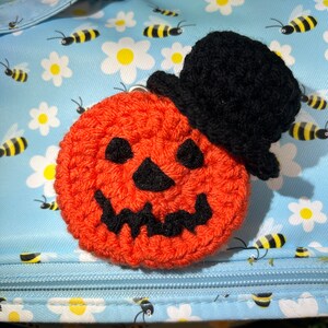Puede incluir: Una calabaza naranja de crochet con un sombrero de copa negro. La calabaza tiene una cara de calabaza tallada con ojos, nariz y boca negros.