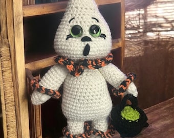 Puede incluir: Muñeca fantasma de ganchillo blanca, con ojos grandes y una expresión de sorpresa. Tiene detalles en negro y naranja, incluyendo una bufanda y envolturas en brazos y piernas. La muñeca sostiene una bolsa de truco o trato negra con relleno verde. El texto en la imagen dice "Carnival Ghost By: A Stitch Of Mine Designs."