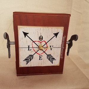 Puede incluir: Un reloj de madera con un corazón rojo y la palabra "LOVE" en el centro. El reloj está rodeado de cuatro flechas y tiene una esfera de reloj de color dorado. El reloj está montado sobre una base de madera con dos asas en forma de pistola.