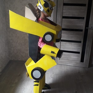 Puede incluir: Un ni&ntilde;o vistiendo un disfraz de robot de cart&oacute;n amarillo y negro. El disfraz tiene una cabeza grande con una m&aacute;scara facial negra y un casco amarillo. El disfraz tiene un ala grande en la espalda y un brazo grande en el lado derecho.