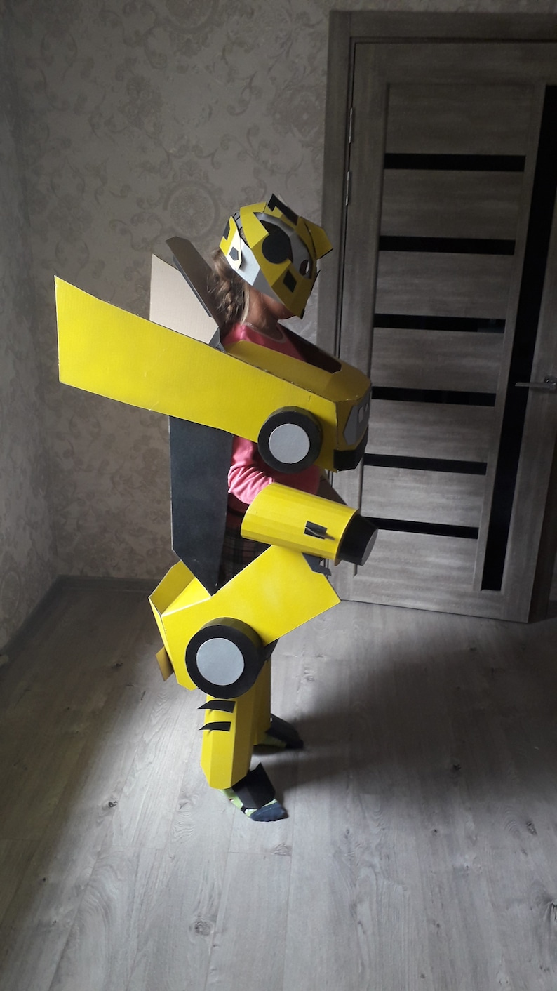 Puede incluir: Un ni&ntilde;o vistiendo un disfraz de robot de cart&oacute;n amarillo y negro. El disfraz tiene una cabeza grande con una m&aacute;scara facial negra y un casco amarillo. El disfraz tiene un ala grande en la espalda y un brazo grande en el lado derecho.
