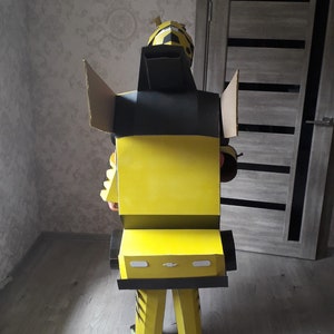 Puede incluir: Un disfraz de cart&oacute;n de Bumblebee de la franquicia Transformers. El disfraz es amarillo y negro con una cabeza negra y un cuerpo amarillo. El disfraz tiene forma de robot y tiene un dise&ntilde;o de coche en la parte delantera.