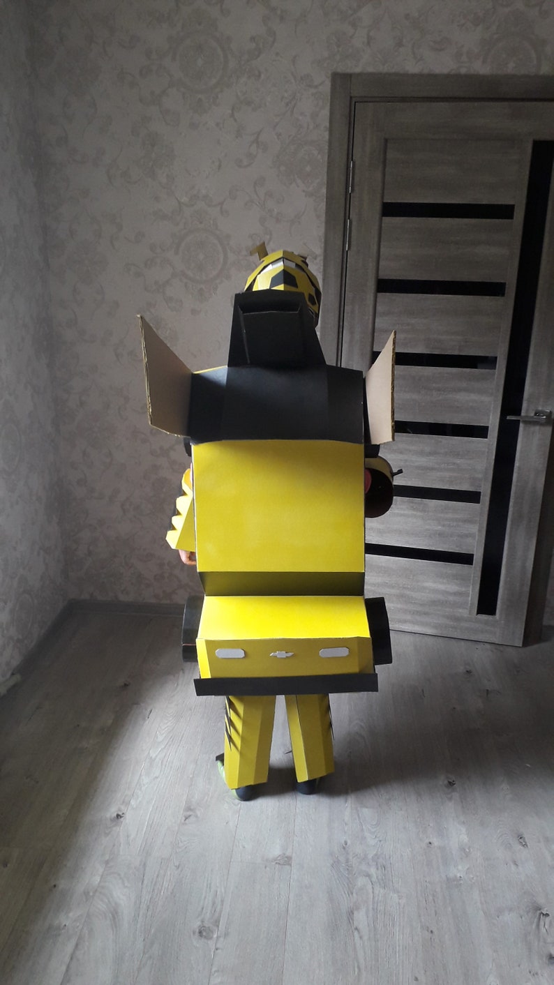 Puede incluir: Un disfraz de cart&oacute;n de Bumblebee de la franquicia Transformers. El disfraz es amarillo y negro con una cabeza negra y un cuerpo amarillo. El disfraz tiene forma de robot y tiene un dise&ntilde;o de coche en la parte delantera.