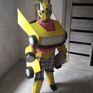 Puede incluir: Un ni&ntilde;o lleva un disfraz de Bumblebee de cart&oacute;n amarillo y negro. El disfraz est&aacute; hecho de cart&oacute;n e incluye una pieza para la cabeza, brazos, piernas y una pieza para el pecho. El ni&ntilde;o est&aacute; de pie frente a una puerta de madera.