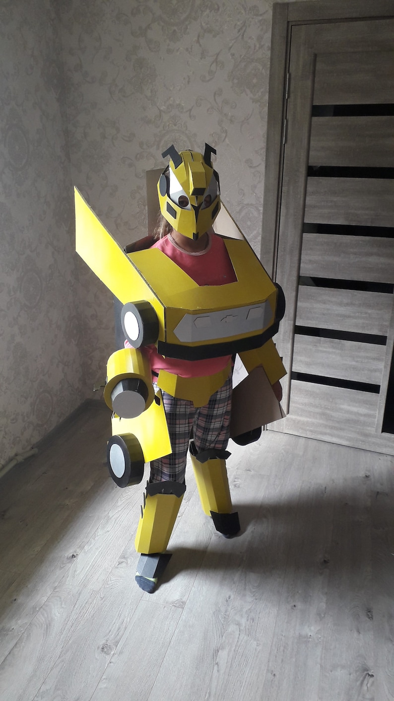 Puede incluir: Un ni&ntilde;o lleva un disfraz de Bumblebee de cart&oacute;n amarillo y negro. El disfraz est&aacute; hecho de cart&oacute;n e incluye una pieza para la cabeza, brazos, piernas y una pieza para el pecho. El ni&ntilde;o est&aacute; de pie frente a una puerta de madera.