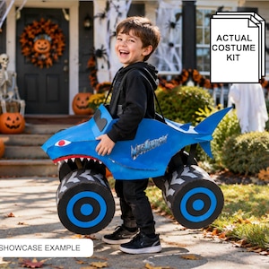 Op de afbeelding: Een blauw monstertruck-kostuum met een haaiendesign, een rode mond, scherpe witte tanden en grote zwart-blauwe wielen. Op de zijkant van de truck staat de tekst Megalodon. De afbeelding bevat de tekstlabels Actual Costume Kit en Showcase Example.