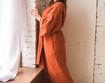 May include: A woman wearing a long, rust-colored robe stands near a window with white curtains. The robe has a tie belt and long sleeves. She is barefoot and standing on a wooden floor.