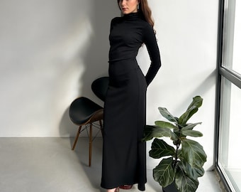 May include: A woman wearing a black turtleneck long-sleeve shirt and a black maxi skirt. She is standing in a room with a white wall and a window. There is a chair and a plant in the room.