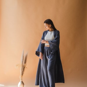 以下が含まれることがあります： 女性が着ているブルーグレーのラウンジウェアセット。セットには、袖の広いロングローブ、クロップドトップ、ワイドレッグパンツが含まれます。背の高い草が入った花瓶と、お揃いのベルトが床にあります。